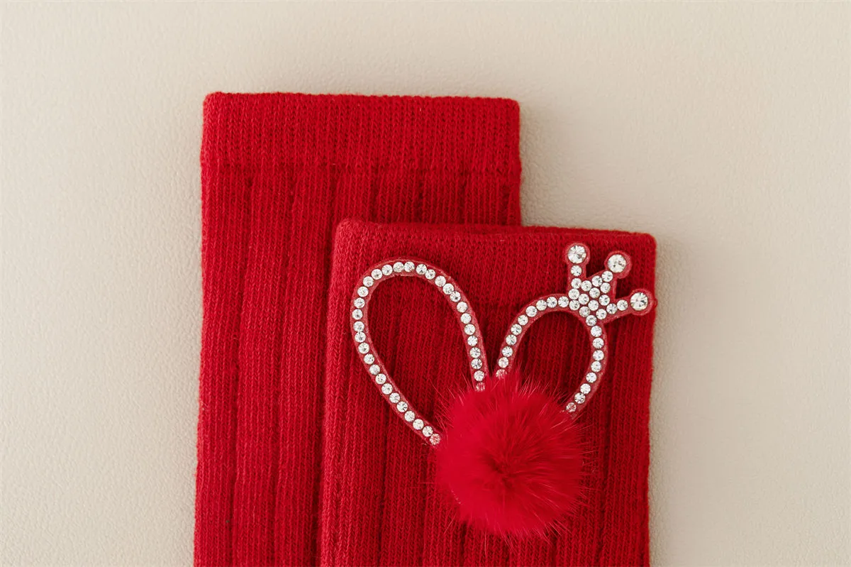 Close-up of red baby socks featuring a rhinestone crown and fluffy pom pom detail.